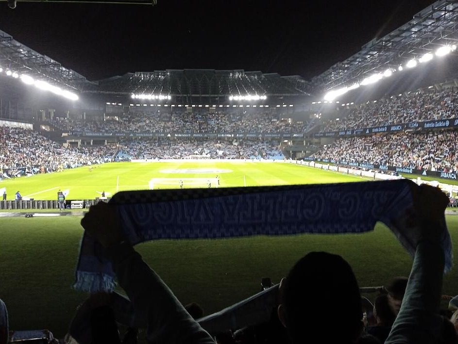 A fan raising a Celta scarf in front of a packed ABANCA Balaídos at a night match