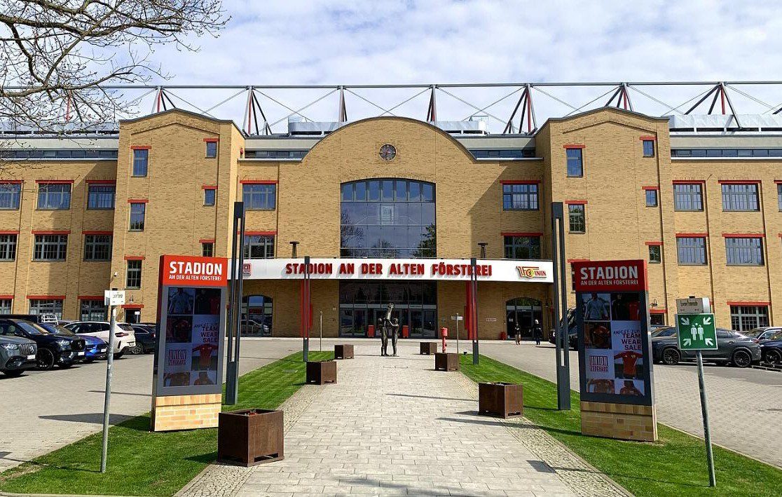 The walkway in front of the main entrance of the Alte Försterei with signboards on both sides and the brick main building behind