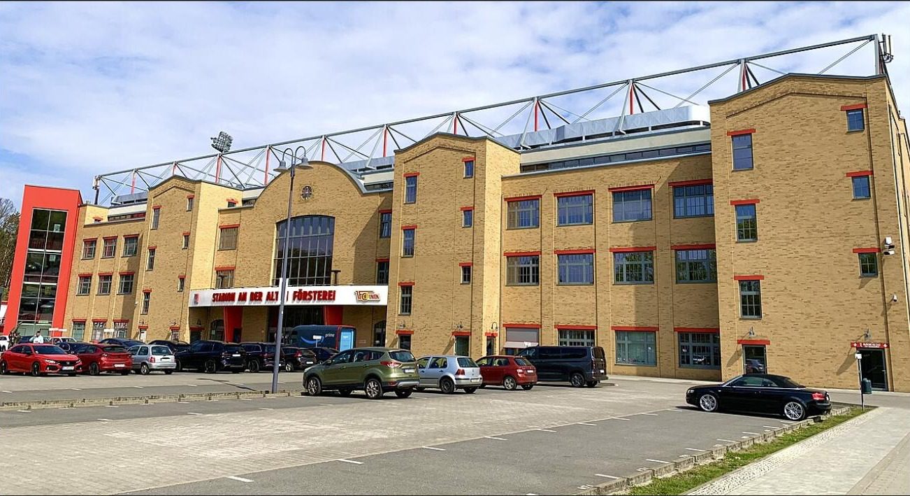 The main stand of the Alte Försterei with a light brick facade and red window frames, and the car park in front