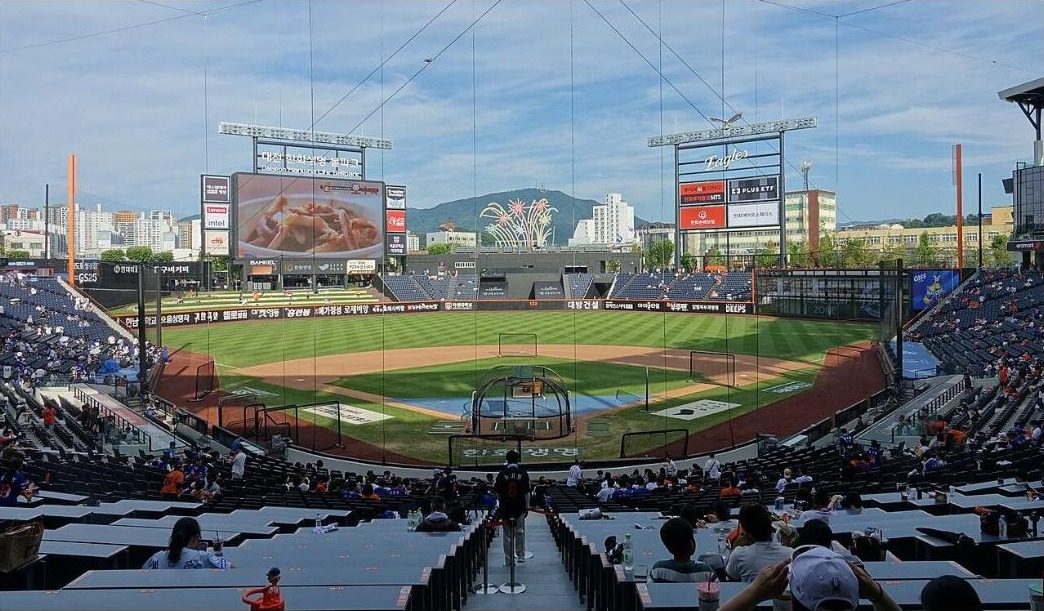 The field and outfield scoreboard of Daejeon Hanwha Life Ballpark seen from the infield stands with fans seated, and a mountain in the distance