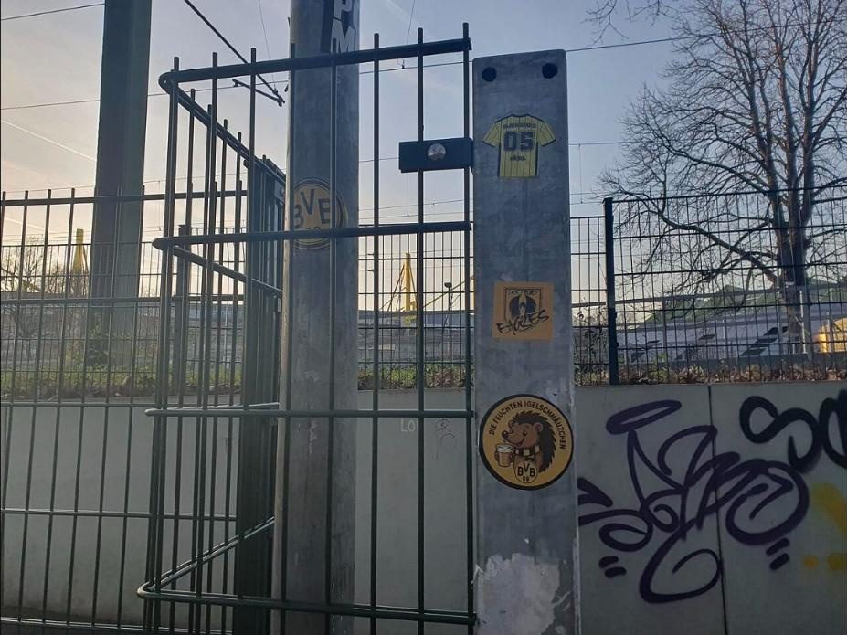 ‘BVB’ 로고 스티커가 붙은 금속 기둥과 울타리, 그 너머로 노란 철골 구조물이 보이는 경전철 웨스트팔렌슈타디온 정류장 진입로