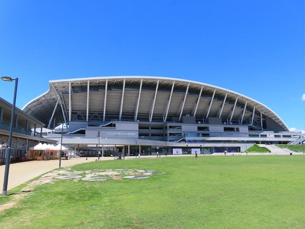 The curved roof and back stand of Edion Peace Wing Hiroshima under a blue sky, seen from the lawn in Chuo Park