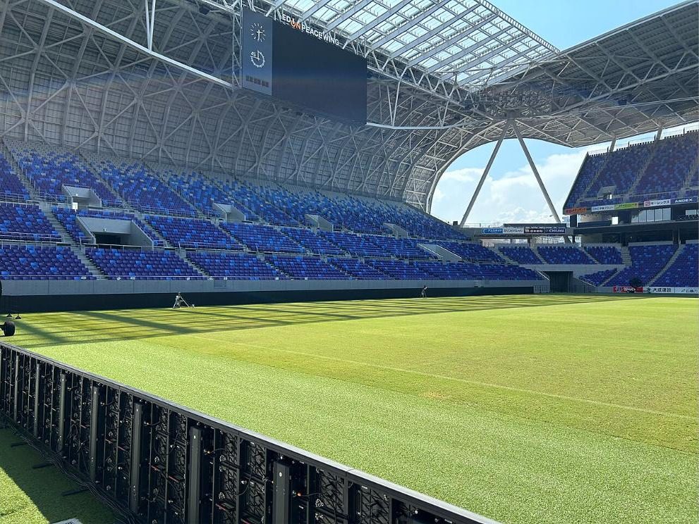 Inside Edion Peace Wing Hiroshima, with blue seats and roof trusses seen from the edge of the pitch