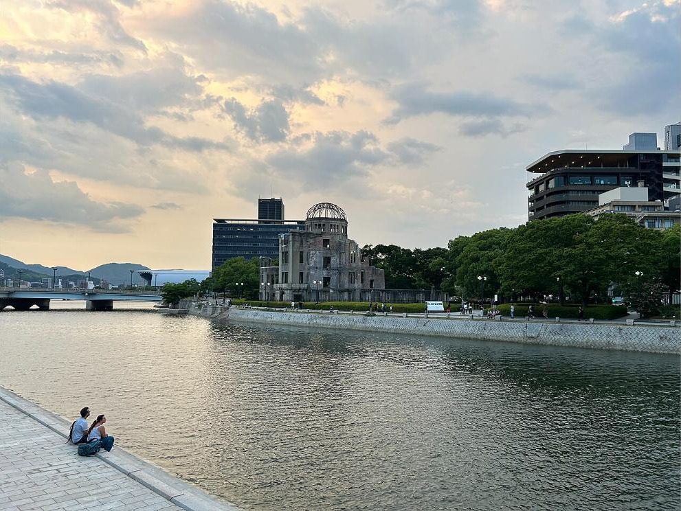 The Atomic Bomb Dome seen across the Motoyasu River at dusk, with two people sitting on the riverbank