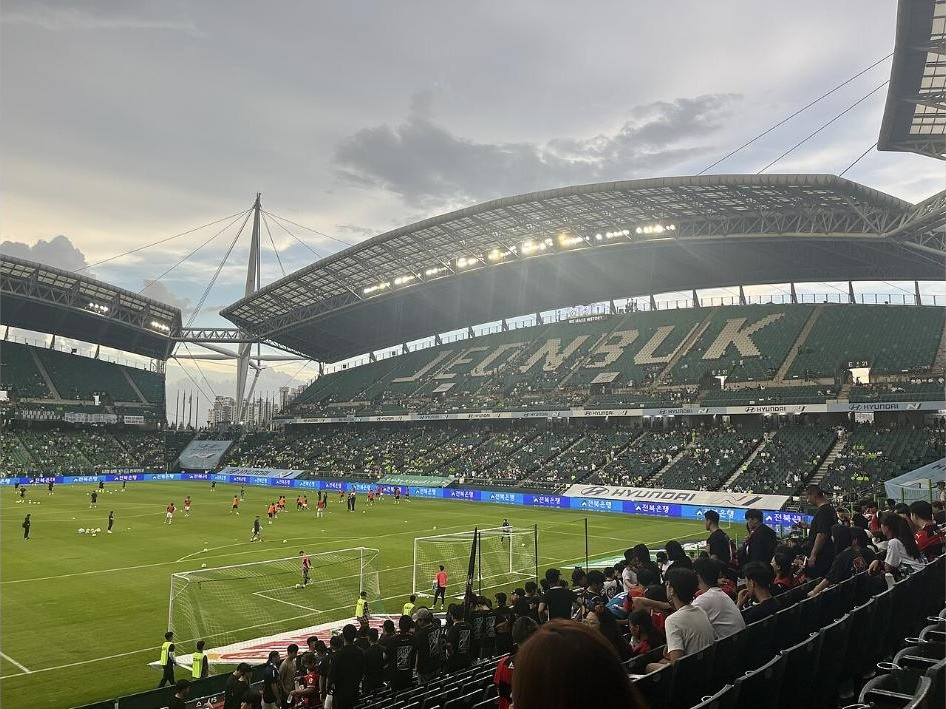 The pitch, the roof and the stands with JEONBUK lettered on them, seen from the away end of Jeonju World Cup Stadium