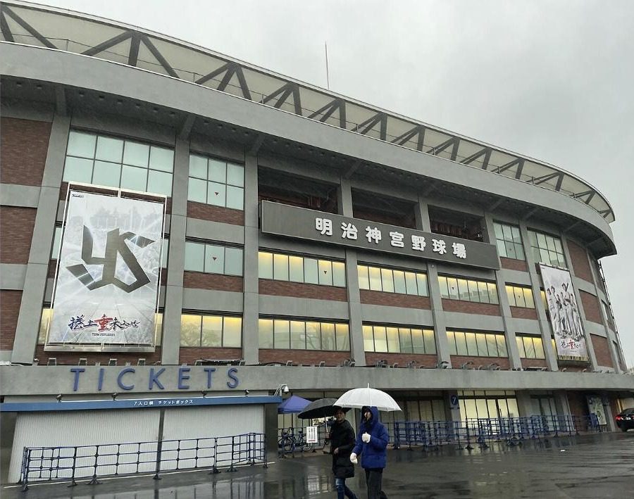 The exterior of Meiji Jingu Stadium on a rainy day with people under umbrellas walking past the Gate 7 ticket box