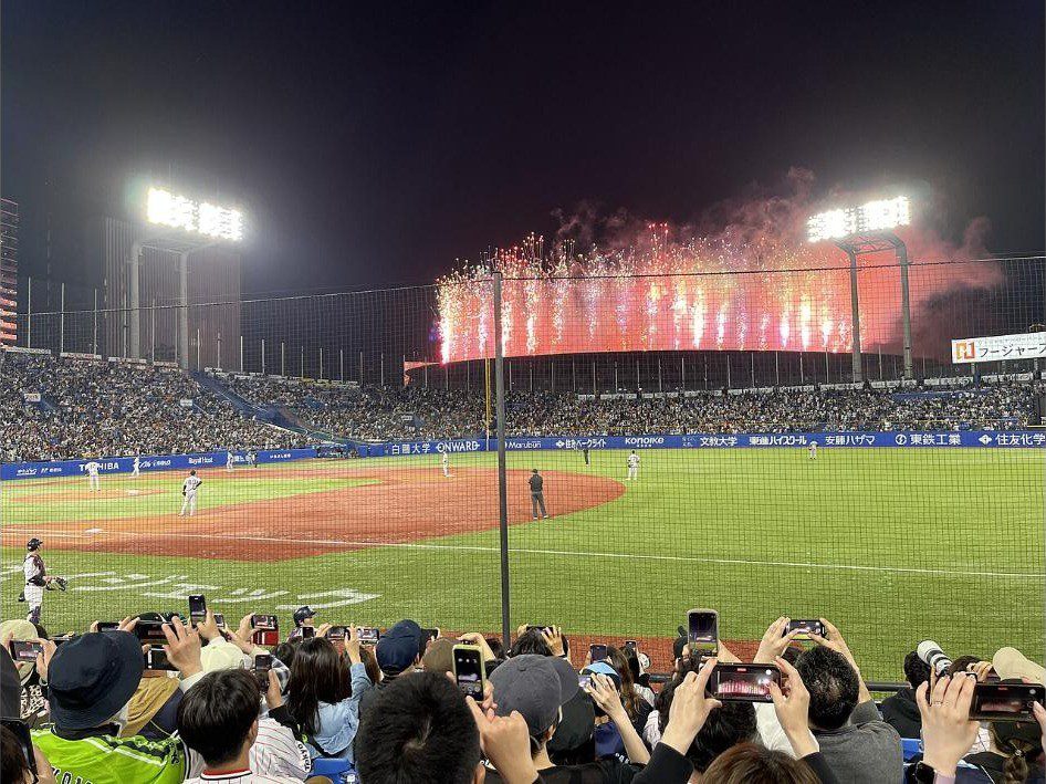 Fireworks bursting behind the outfield at Meiji Jingu Stadium during a night game between the Yakult Swallows and the Yomiuri Giants while fans film with their phones