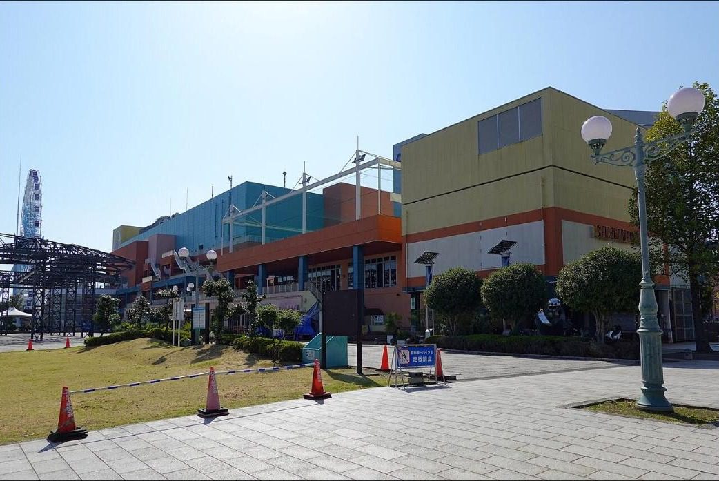 맑은 하늘 아래 주황색과 청록색 건물이 이어진 에스펄스 드림플라자의 외관과 왼쪽 뒤의 대관람차, 앞쪽의 석재 광장과 가로등