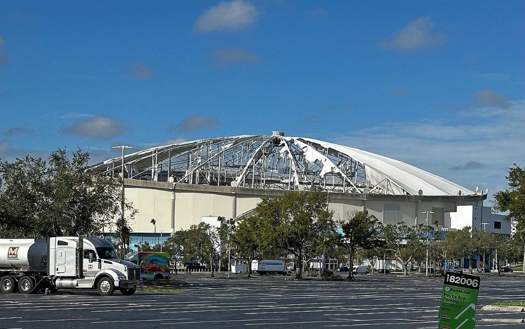 Tropicana Field seen across a parking lot the day after Hurricane Milton, its roof membrane torn away to expose the frame