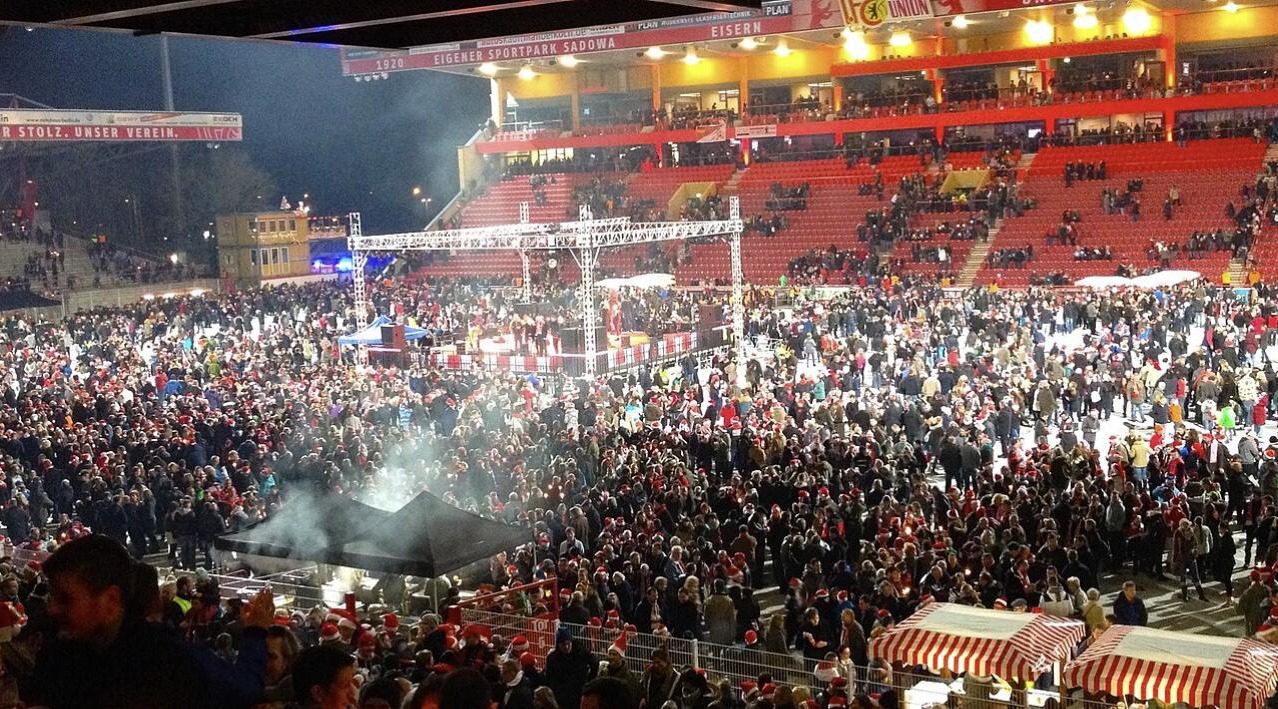 A crowd in red Santa hats packed into the Alte Försterei at night under floodlights, with a temporary stage on the pitch