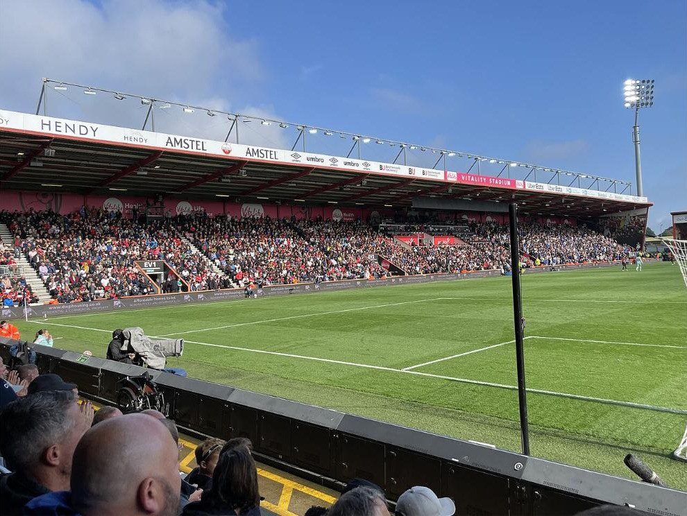 A roofed stand and floodlight pylon at Vitality Stadium, filled with fans under a blue sky